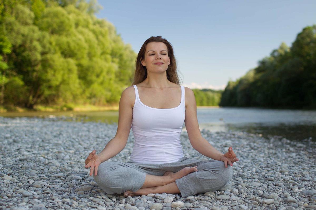 Mujer, vistiendo camisa blanca y pantalón gris, sentada sobre piedras, en posición de meditación, con los ojos cerrados.