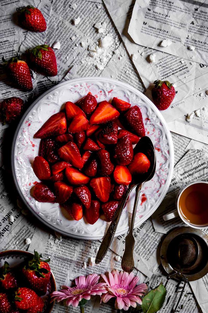 En la mesa cubierta con papel de periódico hay un plato blanco. En ella está la dulce y sabrosa fresa con vinagre. Alrededor hay fresas y entras, cubiertos y una taza de té.