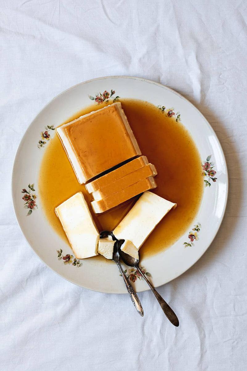 En una mesa cubierta con un mantel blanco hay un plato blanco con el flan de queso sin horno. Ya está cortada y está rodeada con la salsa de caramelo.