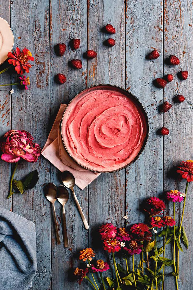 En una mesa de madera azul hay un cuenco azul lleno de helado de frambuesa. El helado se ve cremoso. Alrededor del cuenco hay unas cucharas y flores rojas.