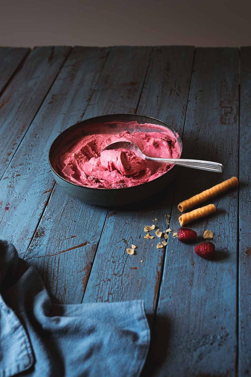 En una mesa de madera azul hay un cuenco azul lleno de helado de frambuesa. En el cuenco hay una cuchara con una bola de helado. Al lado del cuenco hay unas migas de cucurucho y frambuesas frescas. Delante del cuenco hay un paño de cocina azul.