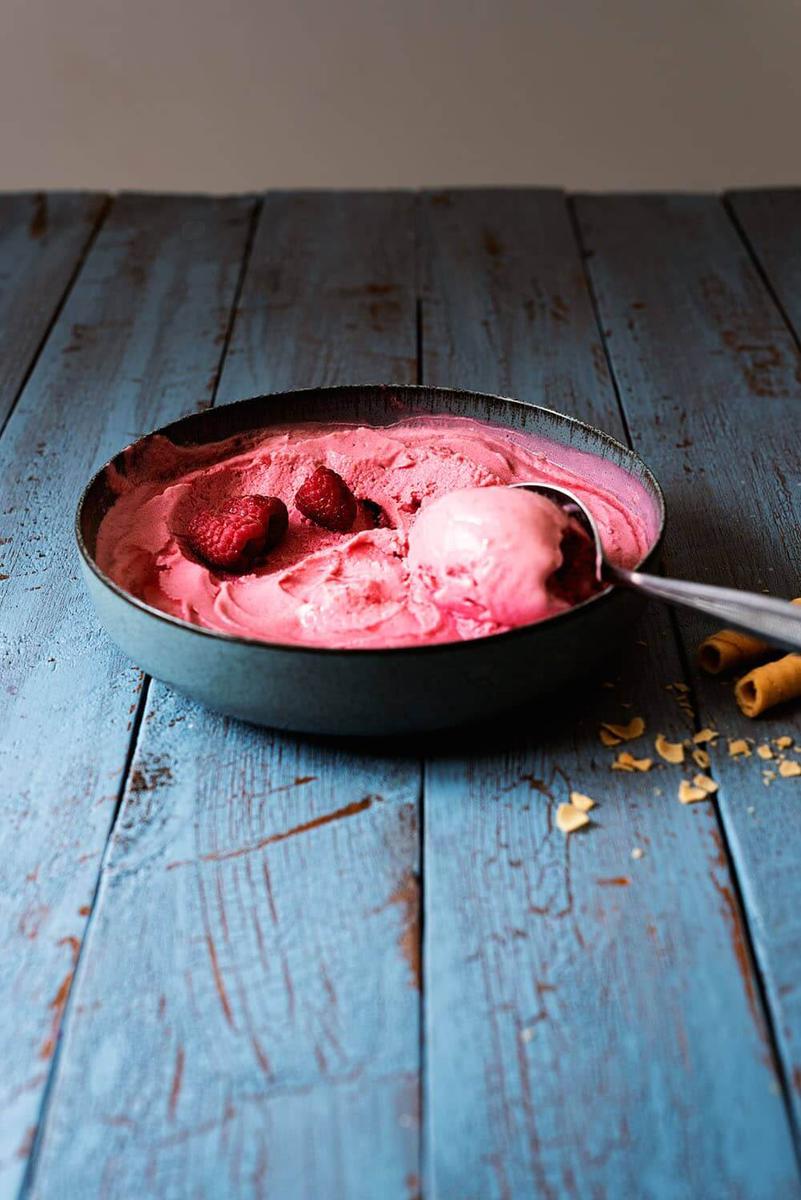 En una mesa de madera azul hay un cuenco azul lleno de helado de frambuesa. En el cuenco hay una cuchara con una bola de helado. Al lado del cuenco hay unas migas de cucurucho.