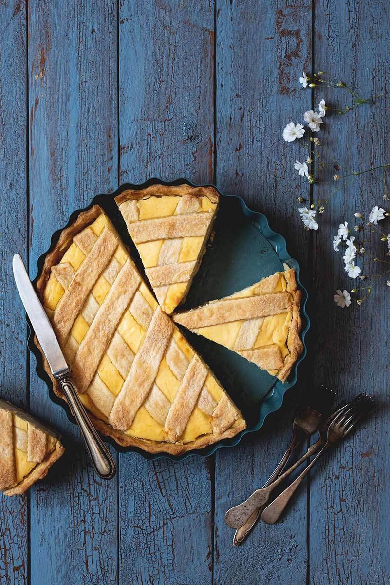 En una superficie azul está la crostata de ricotta. Ya está cortada. Alrededor hay un cuchillo, un par de tenedores y flores blancas.