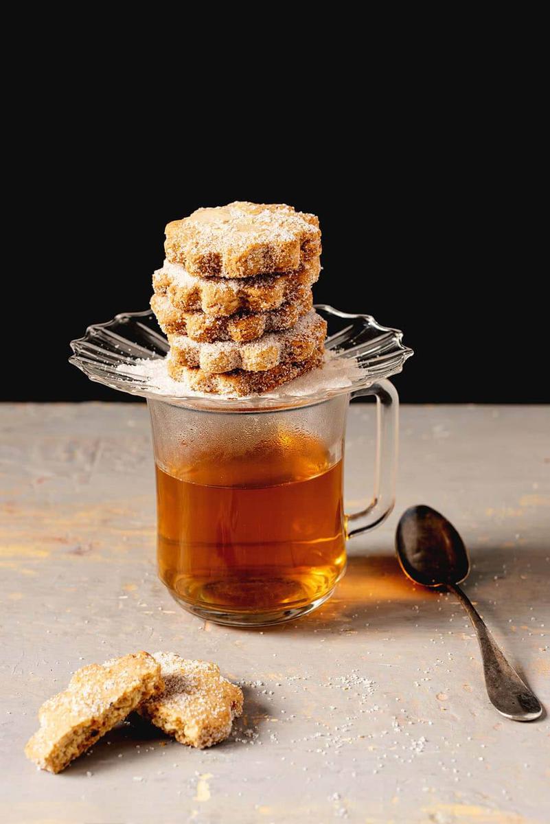 En una superficie gris está un vaso transparente lleno de té caliente. Encima de la taza hay un plato y encima hay varios mantecados manchegos navideños de vino blanco puestos encima una y otra.