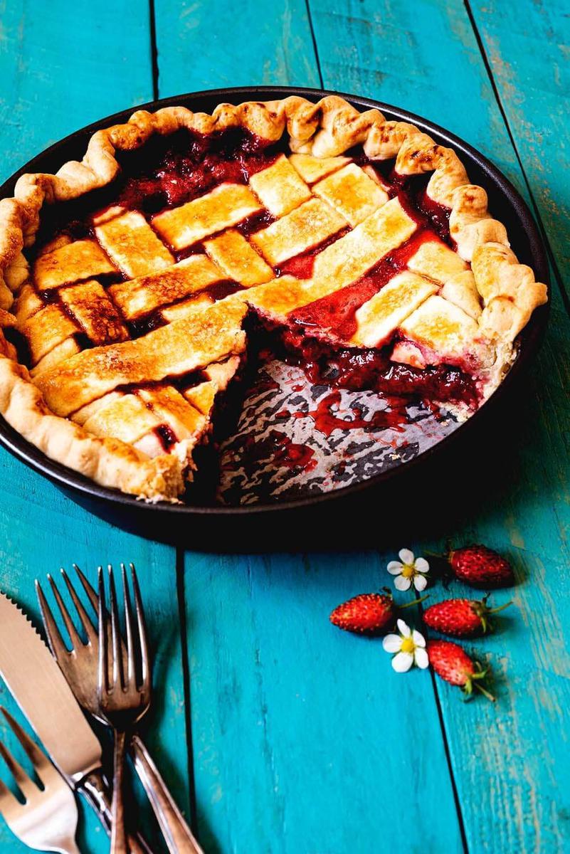 En una mesa azul está el pie de fresa. Al lado hay unas fresas frescas y flor de fresa y cubiertos. El pie está cortado y se ve muy jugoso.