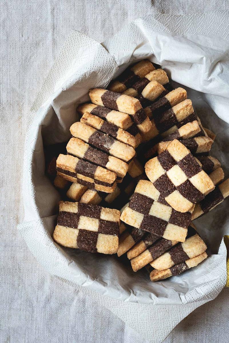 Una mesa está cubierta con un mantel blanco. Encima hay una caja metálica llena de galletas de ajedrez.