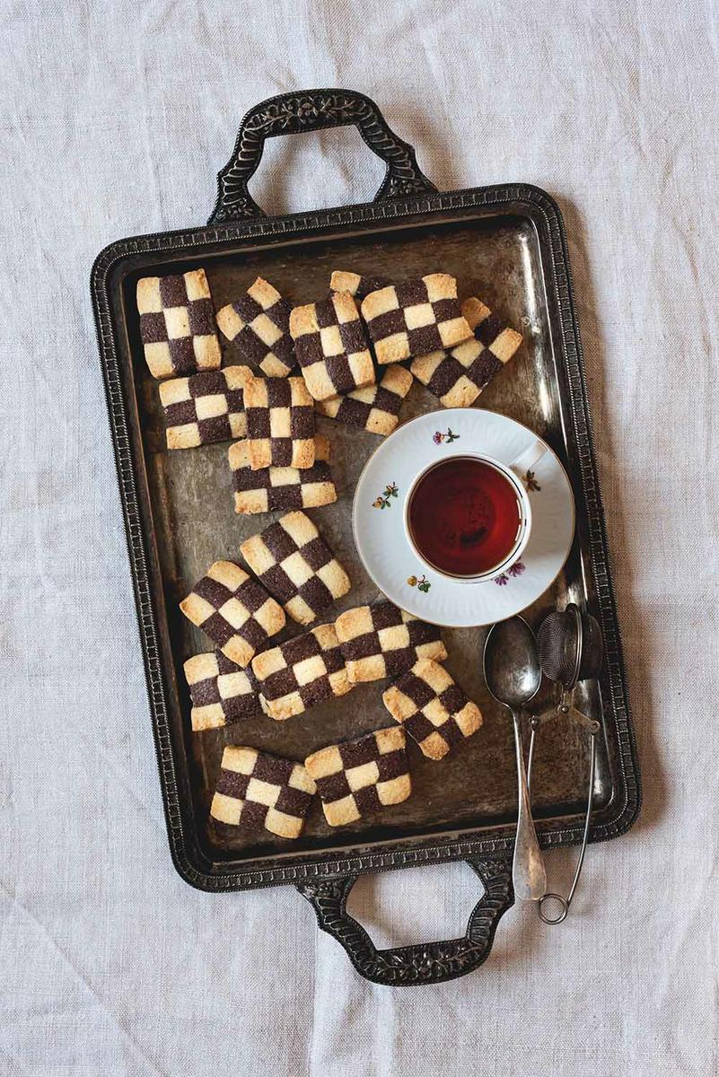 Una mesa está cubierta con un mantel blanco. Encima hay una bandeja metálica. En la bandeja hay galletas damero y una taza de té.