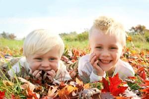 A portrait of two, happy, smiling young children, playing outside in a pile of red and yellow fallen Maple Tree leaves on an Autumn day.