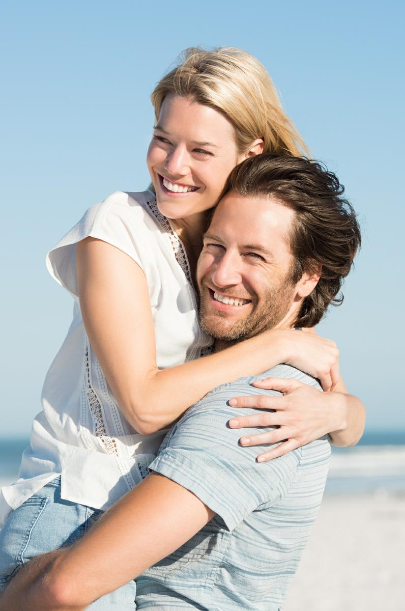 Portrait of a happy boyfriend embracing girlfriend at beach. Handsome man picking up and hugging his girlfriend at sea shore on a sunny day. Young beautiful woman hugging man in love.