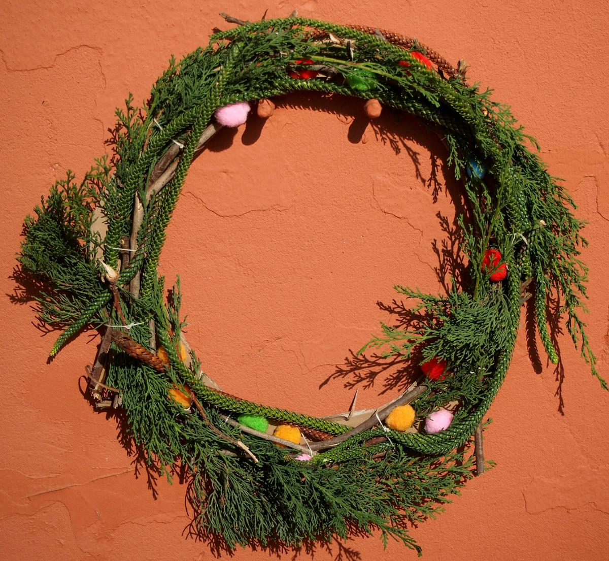 Corona verde hecha de plantas y palos y decorada con pompones. 