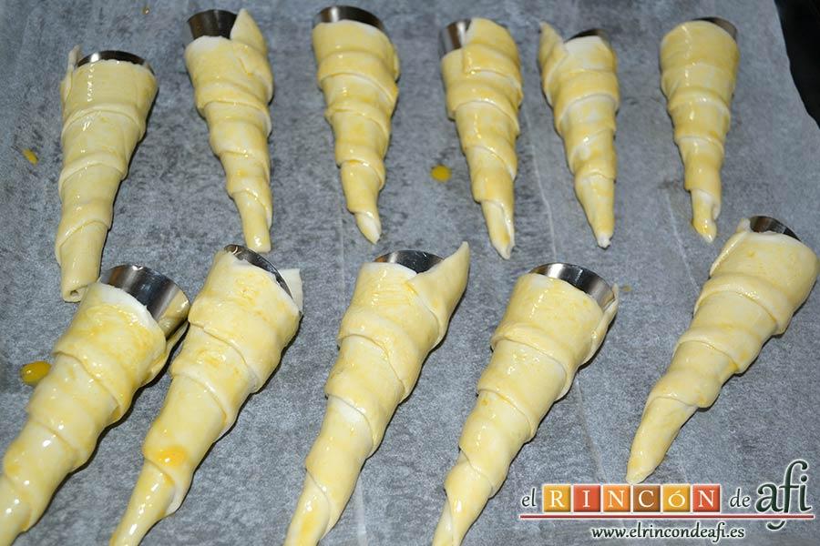 Conos de hojaldre rellenos con crema pastelera, pintar el hojaldre con huevo batido y ponerlos sobre bandeja forrada con papel de horno