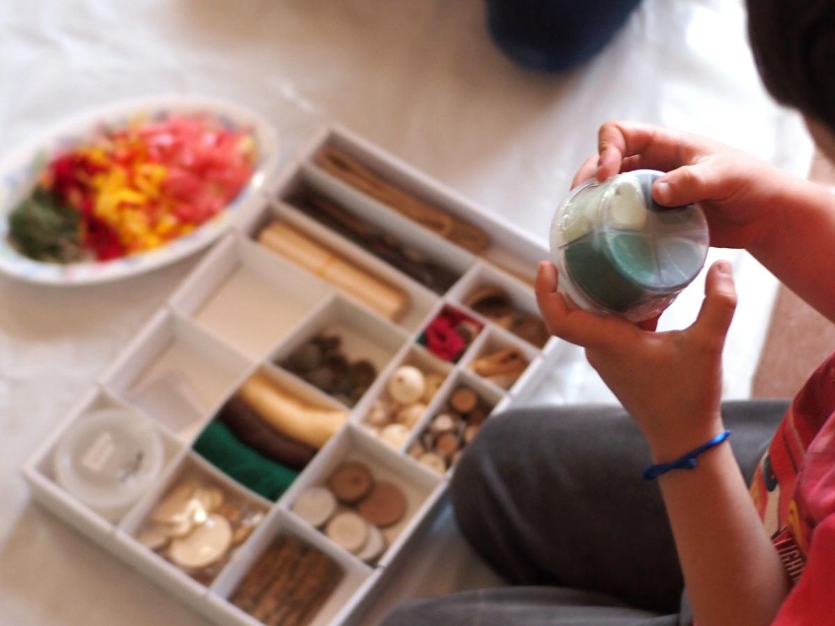 Manos de niño abriendo el bote de pasta de modelar y la caja de piezas sueltas naturales detrás