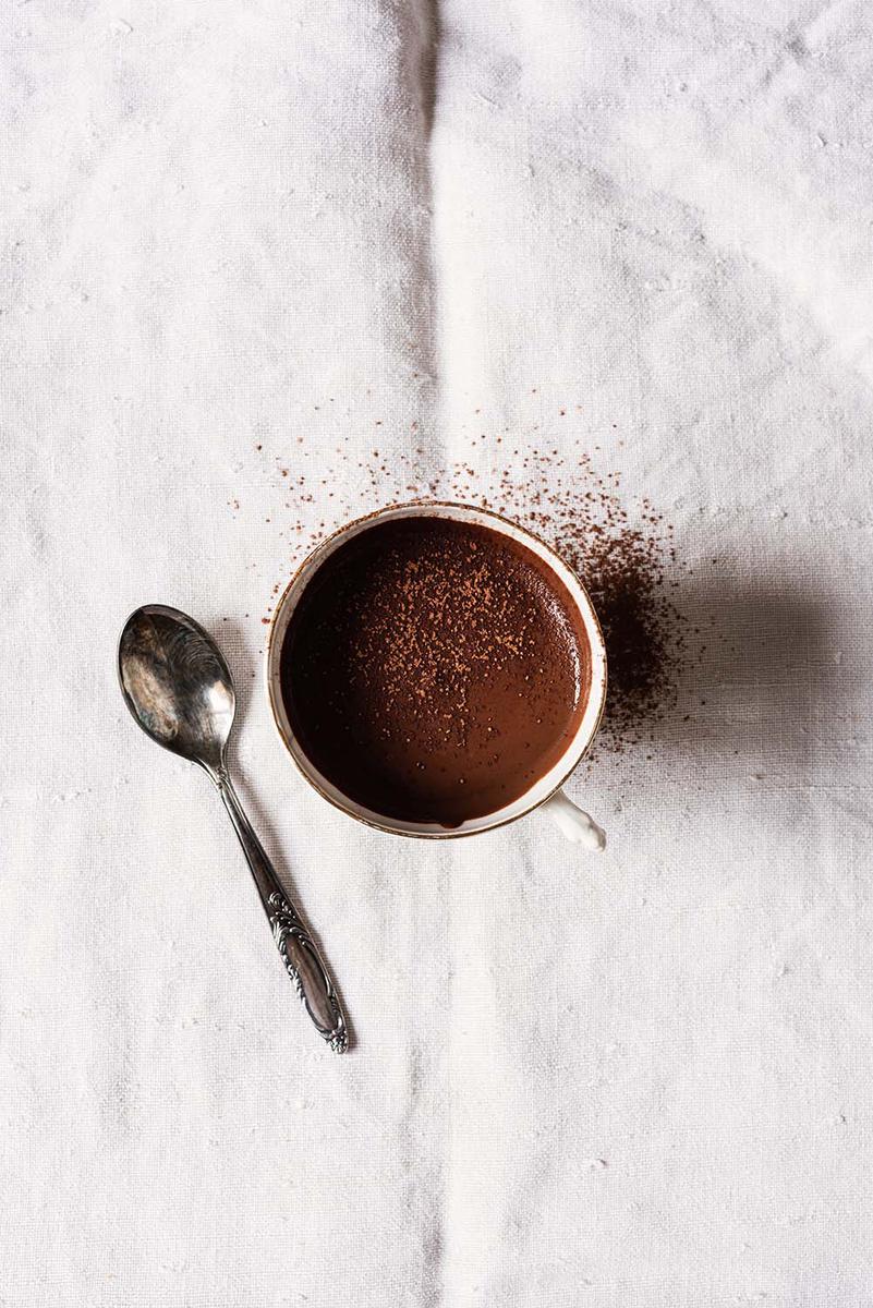 En la mesa cubierta con un mantel blanco, está una taza de chocolate caliente y al lado de la taza está una cuchara vintage.