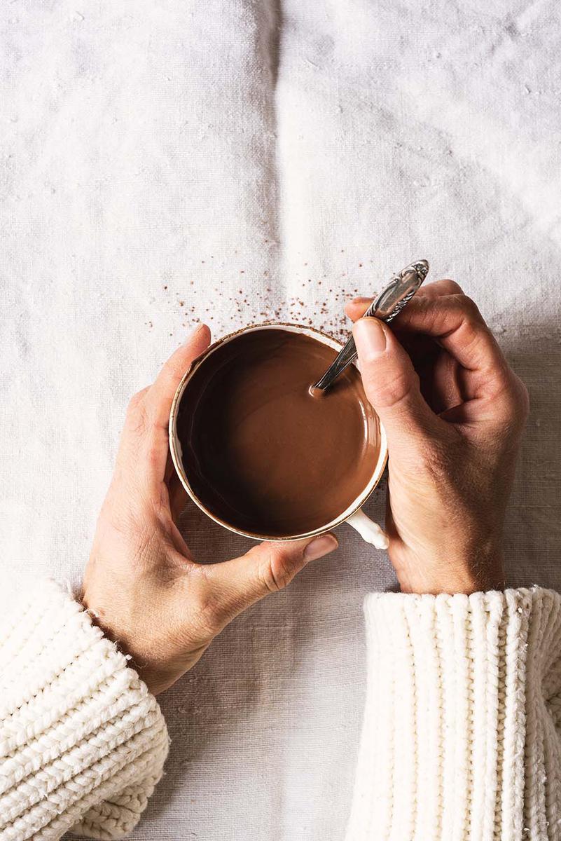 Dos manos están en una mesa cubierta con mantel blanco. Están sujetando una taza de chocolate caliente.