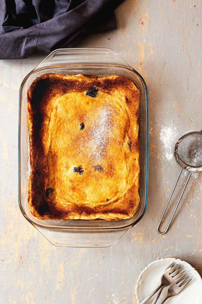 En la mesa gris hay un recipiente donde está el postre Far Bretón sin cortar. En la mesa hay un colador para espolvorear azúcar y un paño de cocina gris.