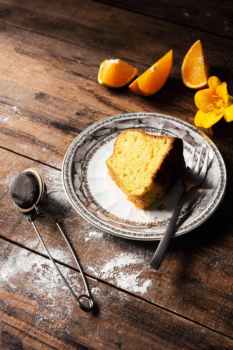 En una mesa de madera está puesto un plato y encima está un trozo de bizcocho de naranja húmedo y esponjoso.