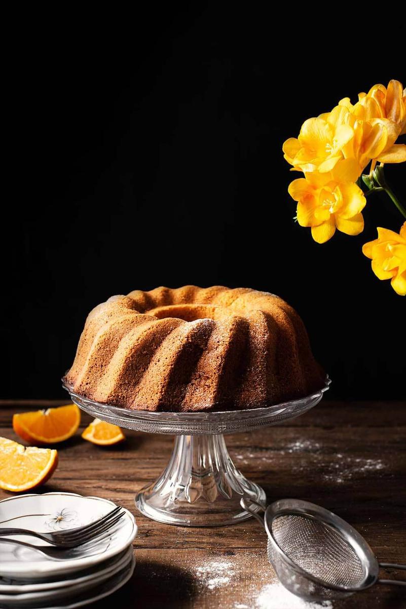 En una mesa madera está puesto un porta tartas cristal y el bizcocho de naranja está encima. En la mesa hay gajos de naranjas y flores amarillas también.