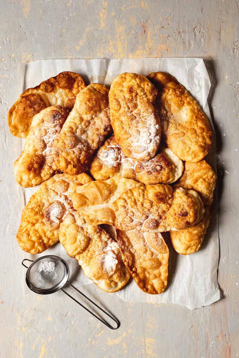 En la mesa está puesto un papel de horno. En ello están amontonadas las Orejas de Carnaval. También se ve un colador para espolvorear con azúcar glas.