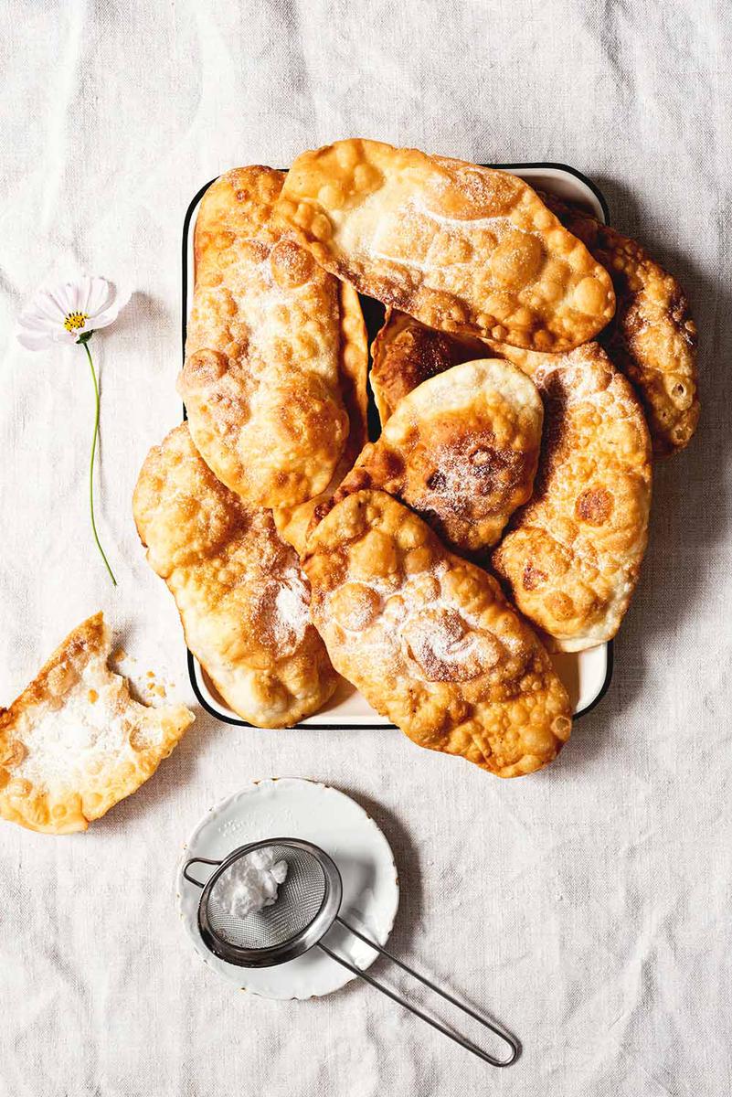 La mesa está cubierta con mantel blanco. Encima hay un plato de hojalata donde están amontonadas las Orejas de Carnaval. También se ve un colador para espolvorear con azúcar glas.