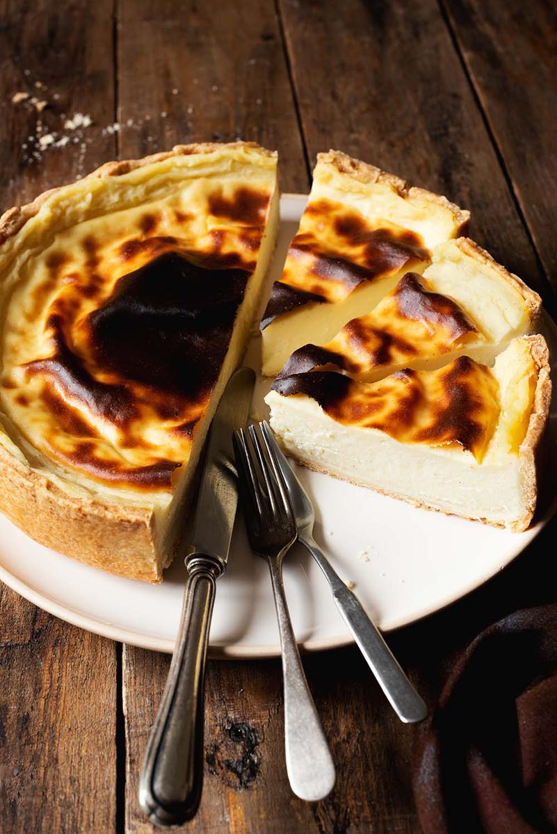 En una mesa de madera está un plato blanco. En este plato está el flan patissier ya cortado. En el plato también hay dos tenedores y un cuchillo. Alrededor del plato hay migas.