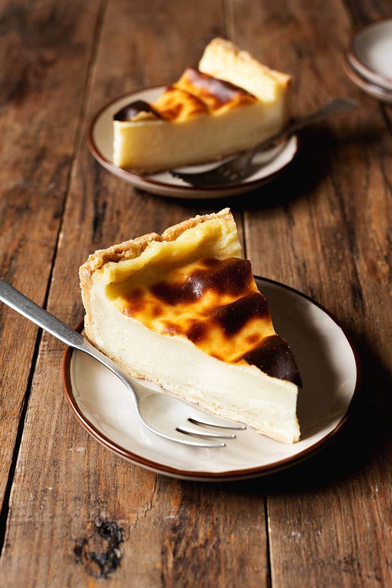 En una mesa de madera está un plato moreno. Encima hay una ración de flan patissier. En este mismo plato hay un tenedor también. Detrás de este trozo de pastel hay otra ración de flan patissier en un plato moreno. En este plato también hay un tenedor.