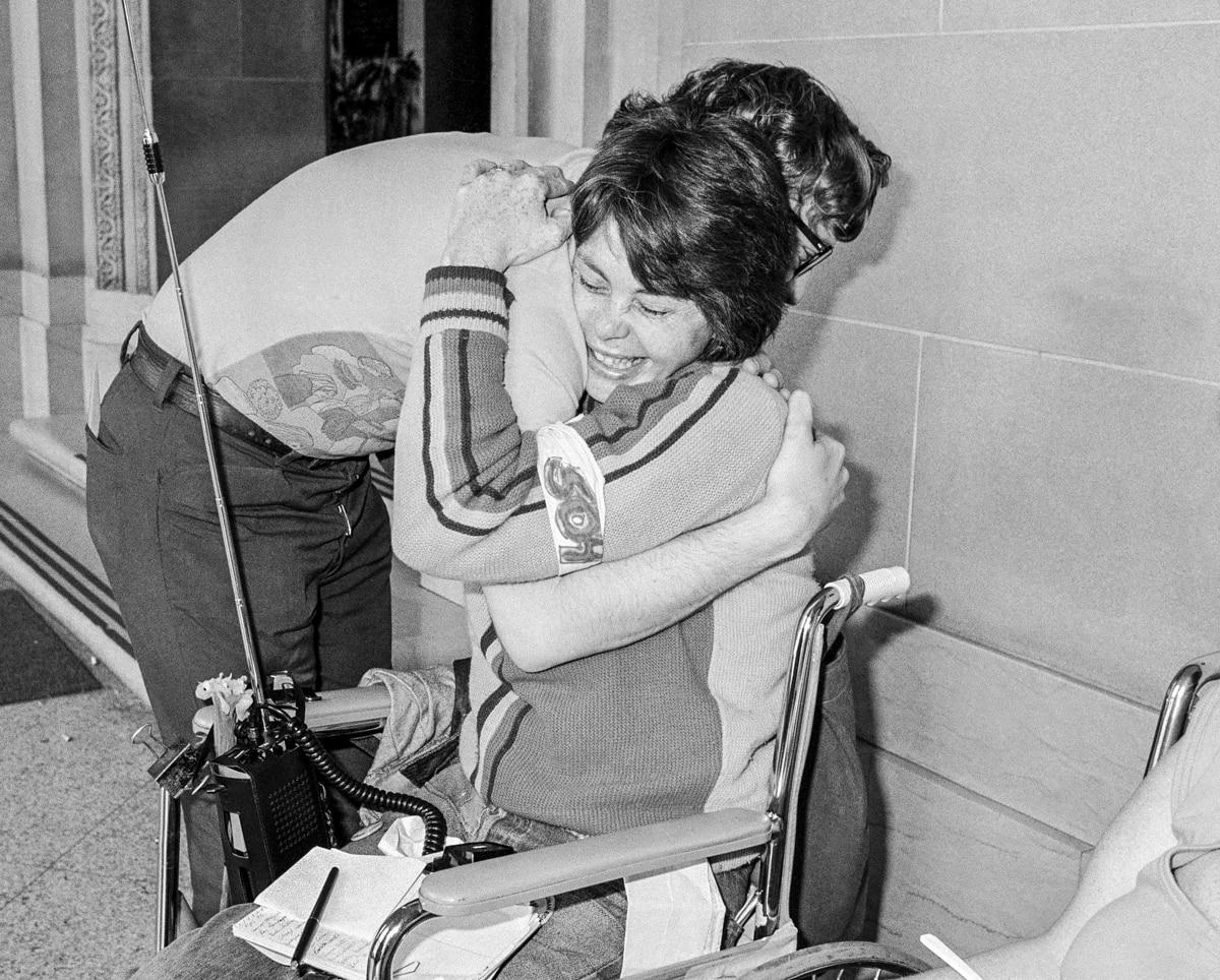 Judy Heumann con una cara feliz y lklena de satisfacción, abrazada a una compañera.