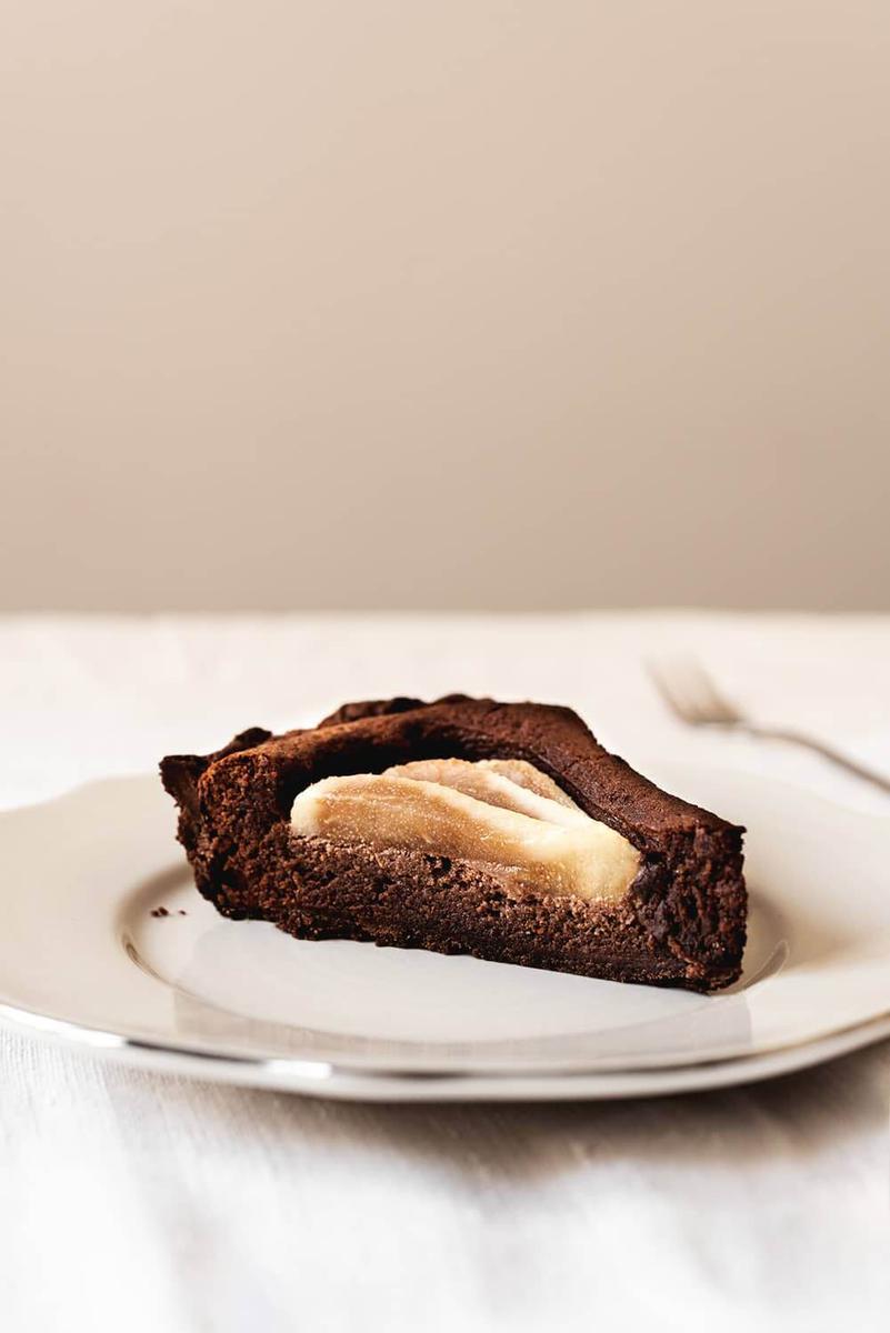 En un plato blanco está un trozo de tarta de peras con chocolate. Se ve un corte bonito de las peras. Detrás del plato hay un tenedor.