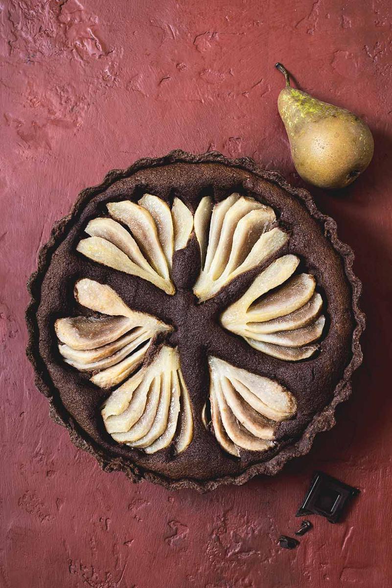 En una mesa de color morada está la tarta de peras con chocolate. A la lado de ella está una pera y un trozo de chocolate.