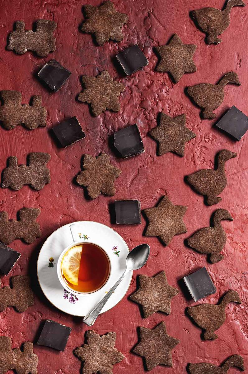 En un mesa de color burdeos hay muchas galletas de cacao puesto. A la derecha hay una taza blanca con té dentro. Al lado de la taza hay una cuchara. En el té hay una media rodaja de limón.