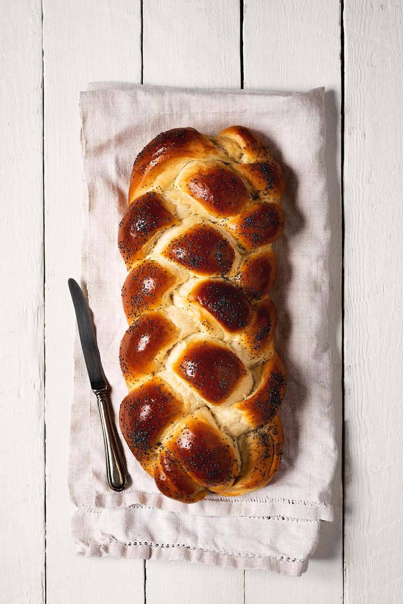 En un paño de cocina blanca está puesto el pan judio sin cortar. Se ve el trenzado de cuatro cabos bonito y la superficie del pan brillante.