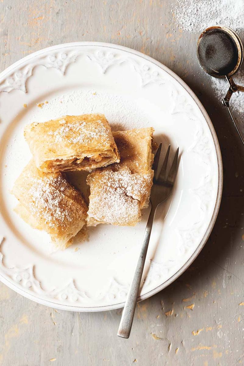 En un plato blanco están tres raciones de strudel de manzana espolvoreados con azúcar glas. En el plato también está puesto un tenedor.