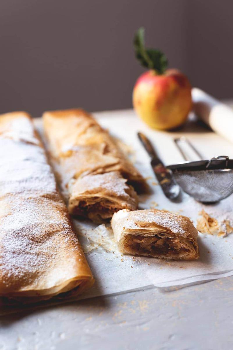 Encima del papel de horno están dos rollos de strudel manzana y dos raciones de strudel manzana, una manzana, un colador pequeño para espolvorear con azúcar glas y un cuchillo.