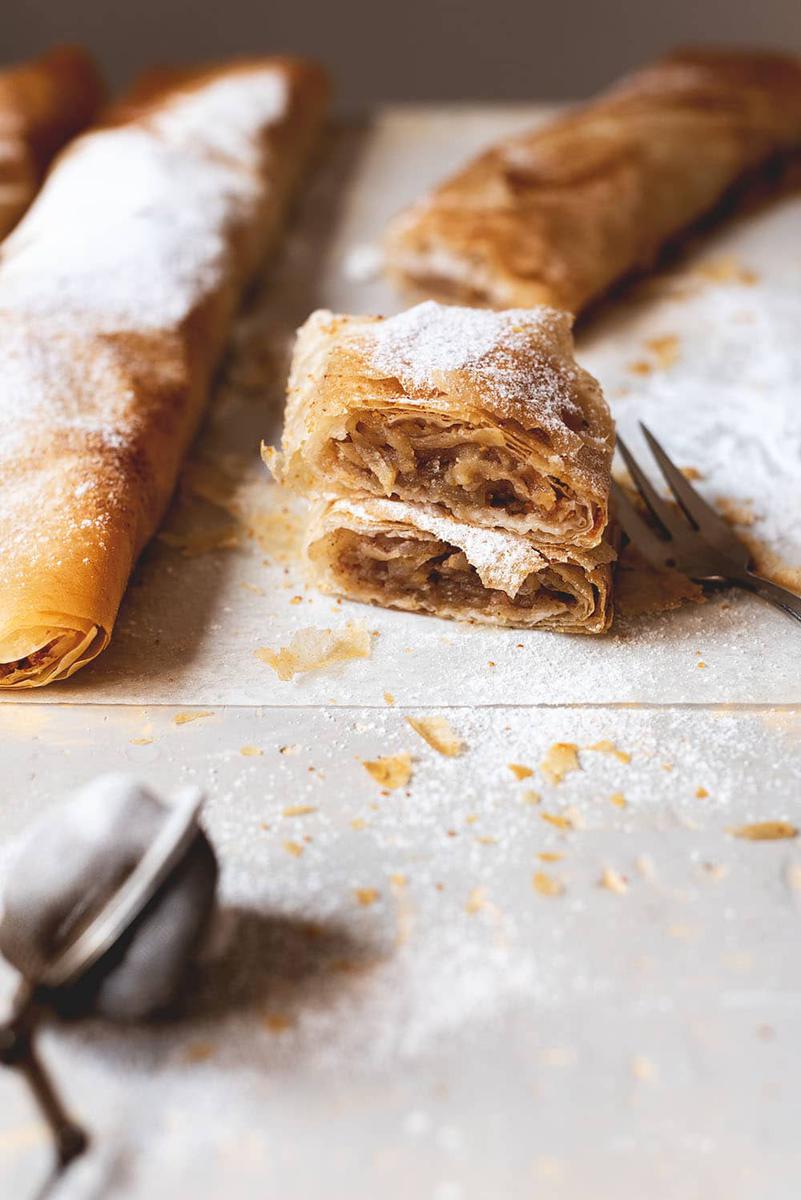 Encima del papel de horno están dos rollos de strudel manzana y dos raciones de strudel manzana puestos encima uno y otra. También se ve una manzana, un colador pequeño para espolvorear con azúcar glas y un cuchillo.