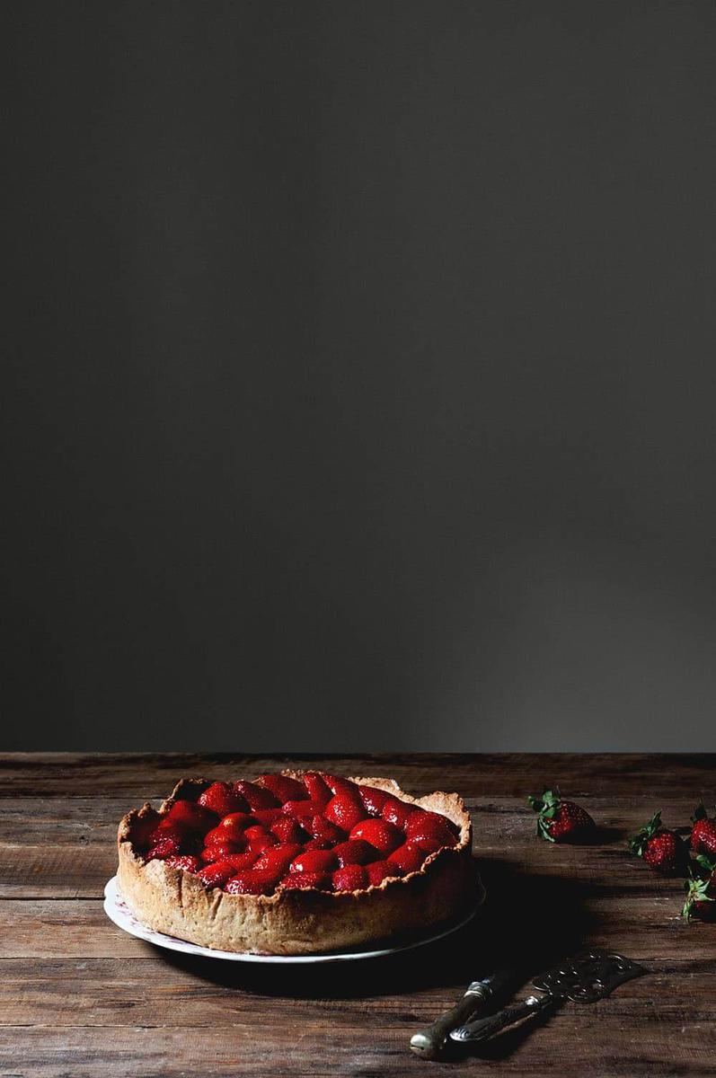 En una mesa de madera está la tarta de fresas con crema y gelatina. Al lado, hay fresas, un cuchillo y una pala de tartas.