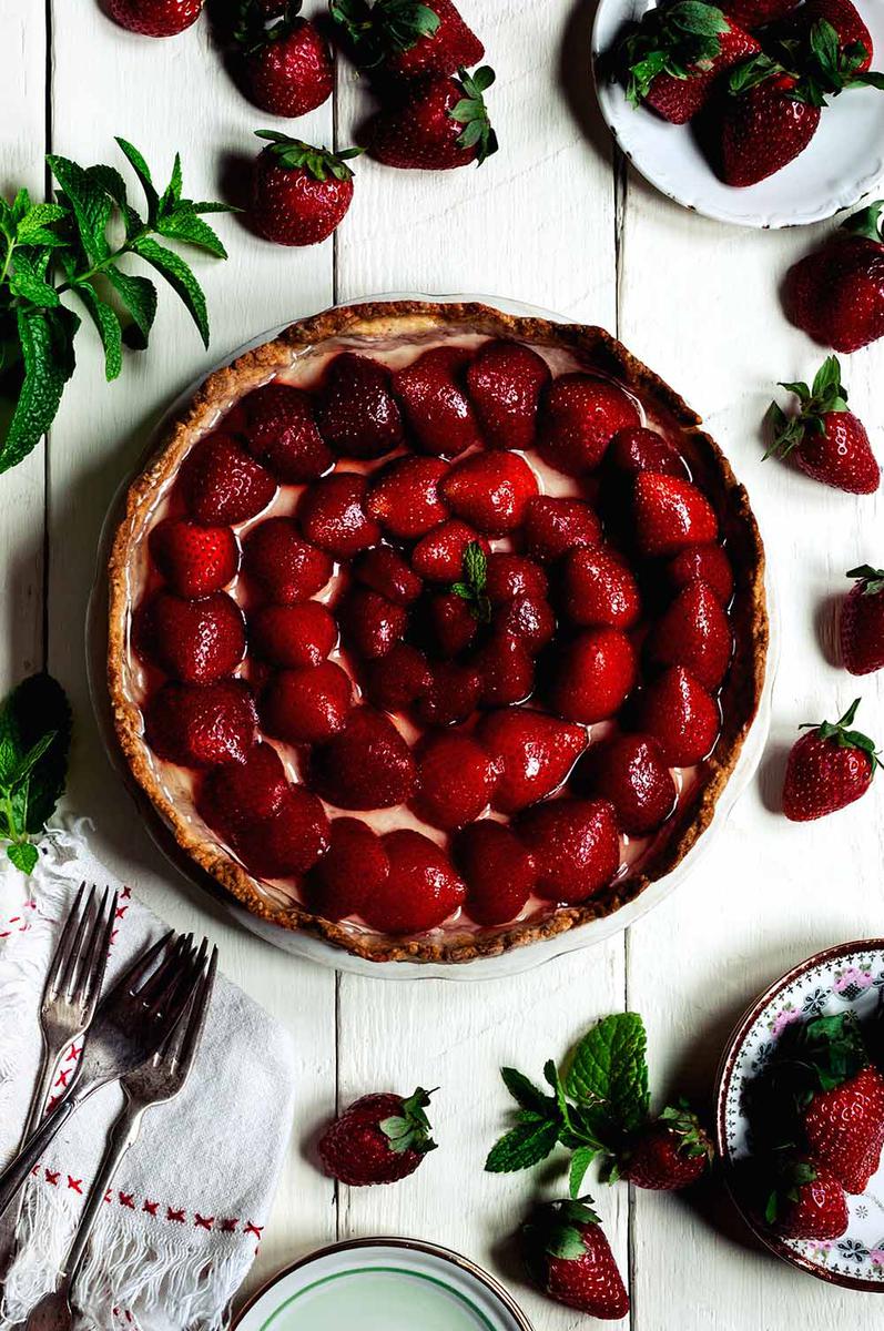 En una mesa blanca está la tarta de fresas con crema. Alrededor de la tarta hay fresas, tenedores, fresas, un paño de cocina blanca y unas hojas de menta.