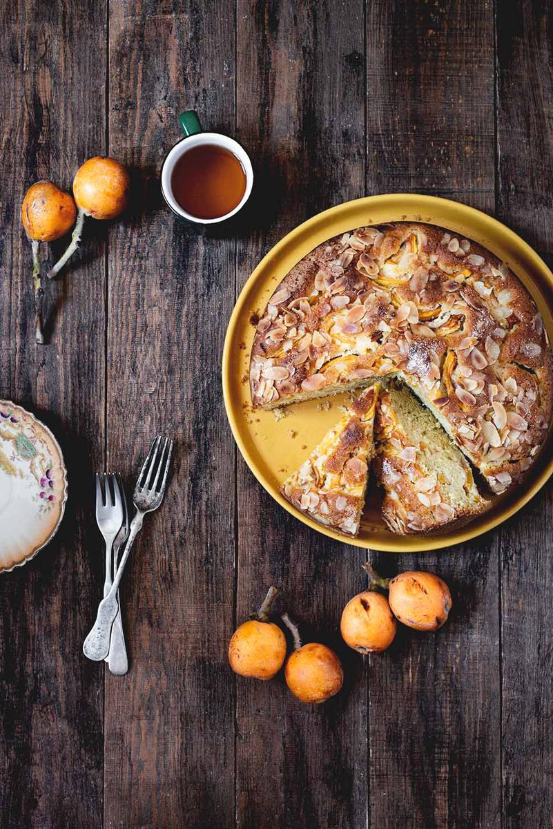 En una mesa de madera esta puesta el bizcocho de nísperos. Alrededor hay cubiertos, una taza de té y unos nísperos frescos. 