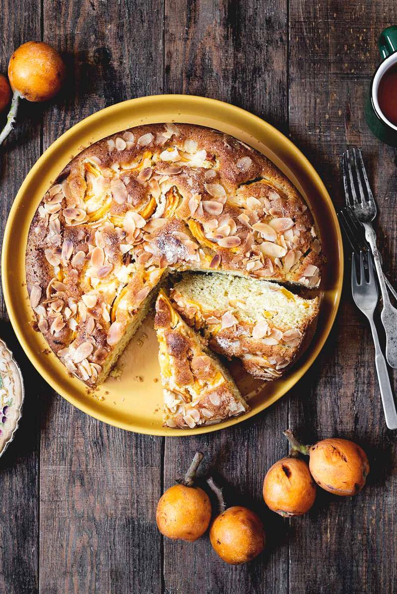 En una mesa de madera está puesta el bizcocho de nísperos. Ya está cortado. Se ve la textura esponjosa. Alrededor hay cubiertos, una taza de té y unos nísperos frescos.