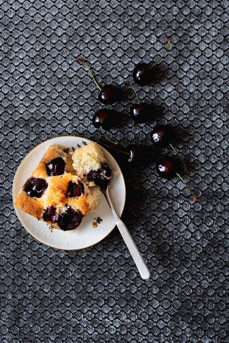 La mesa está cubierta con un mantel gris y encima hay un plato blanco. En ella hay una ración de bizcocho de cerezas. Al lado de este, hay un tenedor. En el mantel hay cerezas frescas.