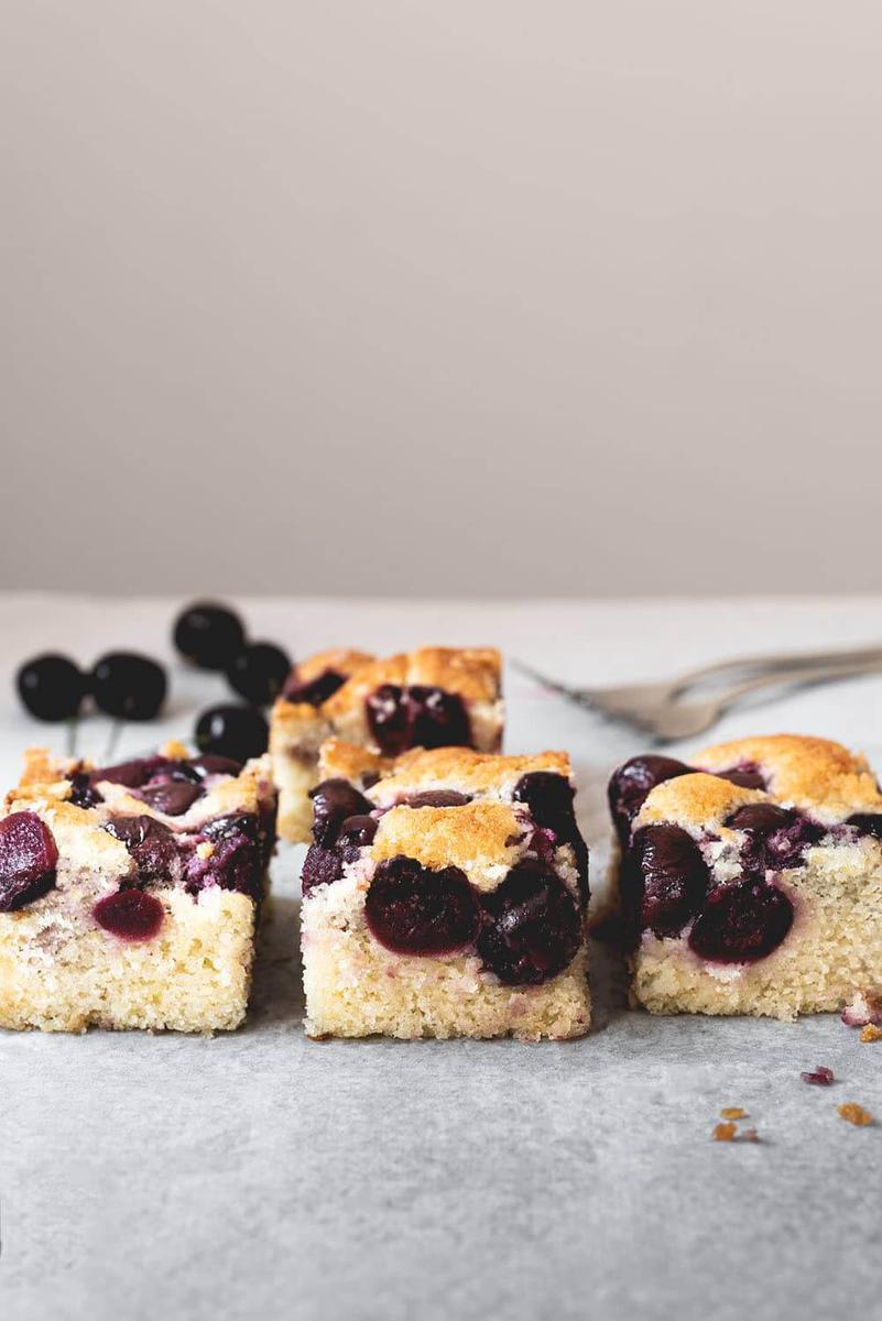 En una mesa gris está el bizcocho de cerezas cortado. Se ve una textura jugosa, también se ven las cerezas. Detrás hay más cerezas frescas y tenedores.