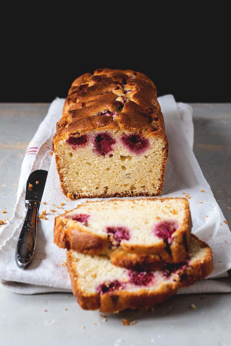 En una mesa gris está puesto un mantel blanco. En el mantel está el bizcocho de frambuesas. Ya está cortado. Se ve una textura esponjosa. Al lado está un cuchillo.