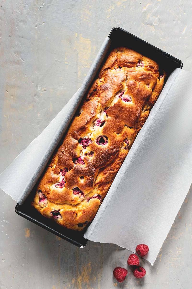 En una mesa de gris hay un molde negro rectangular. En el molde está el bizcocho de frambuesa. Al lado del molde hay varias frambuesas frescas.
