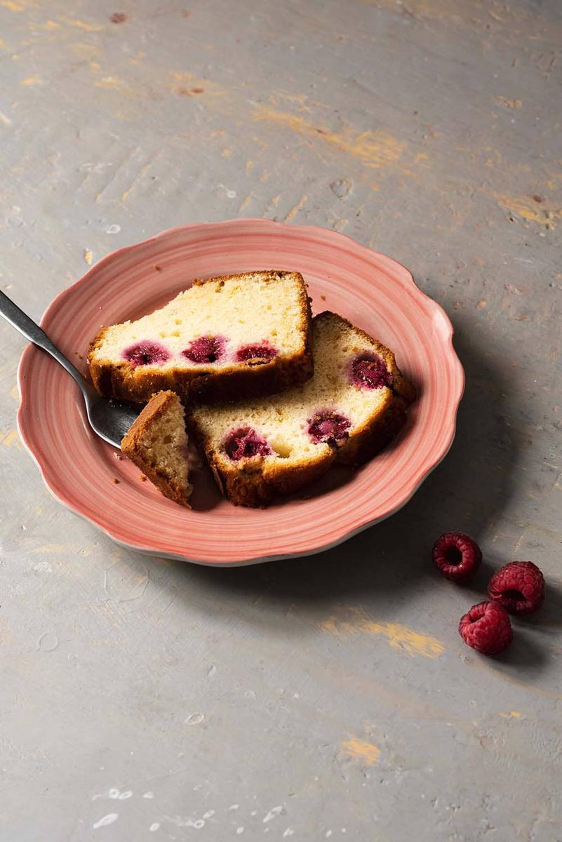 En una mesa de gris hay un plato rosa. En el plato está el bizcocho de frambuesa. Al lado del bizcocho hay un tenedor.