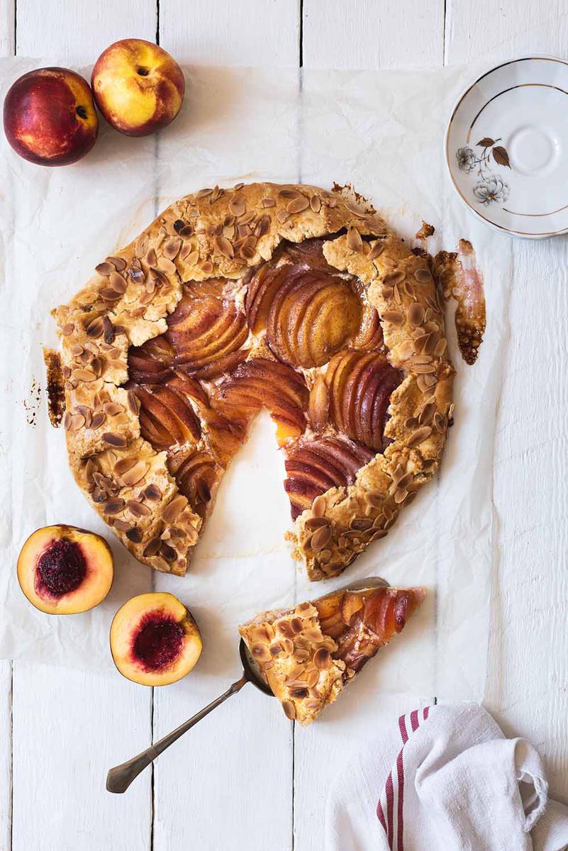 En una mesa de madera blanca está puesto el Galette de nectarina. Alrededor hay un paño de cocina, un plato y varias nectarinas frescas. Y en una pala para tartas hay una ración de galette.