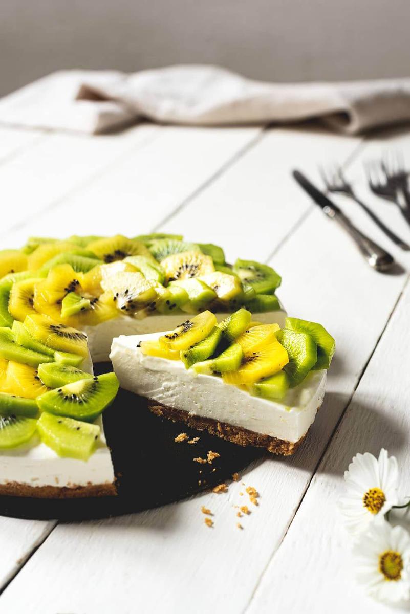 En una mesa de madera blanca está puesta la tarta de kiwi y yogur. Detrás de esta tarta hay cubiertos y un paño de cocina y delante hay una flor. La tarta está cortada y se ve una ración hermosa.