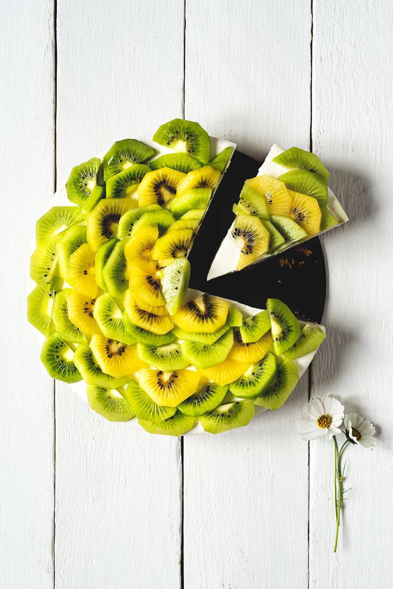 En una mesa de madera blanca está puesta la tarta de kiwi y yogur. La tarta está cortada. Al lado de la tarta hay una flor.