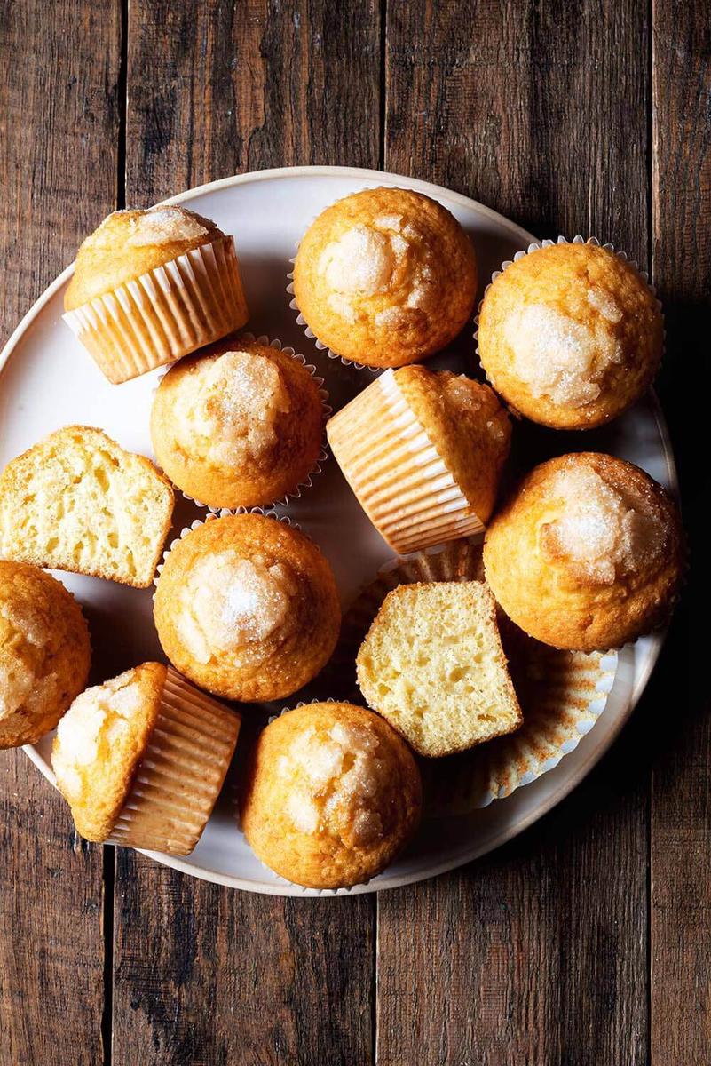 En una mesa de madera, en un plato grande y blanca hay muchas magdalenas. Una de ellas está cortada en la mitad y se ve que esponjoso es.
