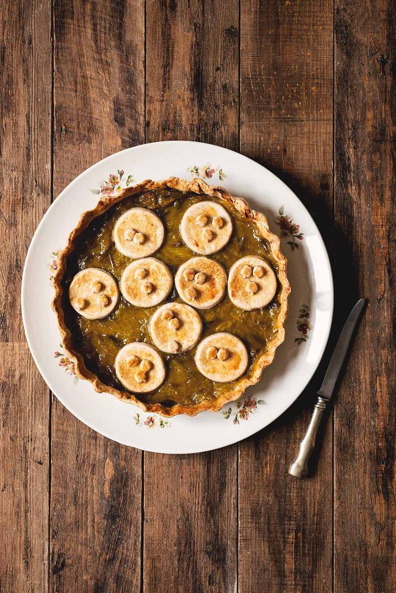 En una mesa de madera hay un plato blanco. En el plato está la tarta de ciruelas Claudia. Está sin cortar. Al lado del plato hay un cuchillo.
