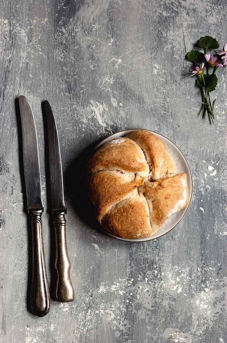 En una mesa gris hay un plato blanco. Encima hay un panecillo Kaiser. Al lado a la izquierda del plato hay dos cuchillos. A la derecha hay unas flores.