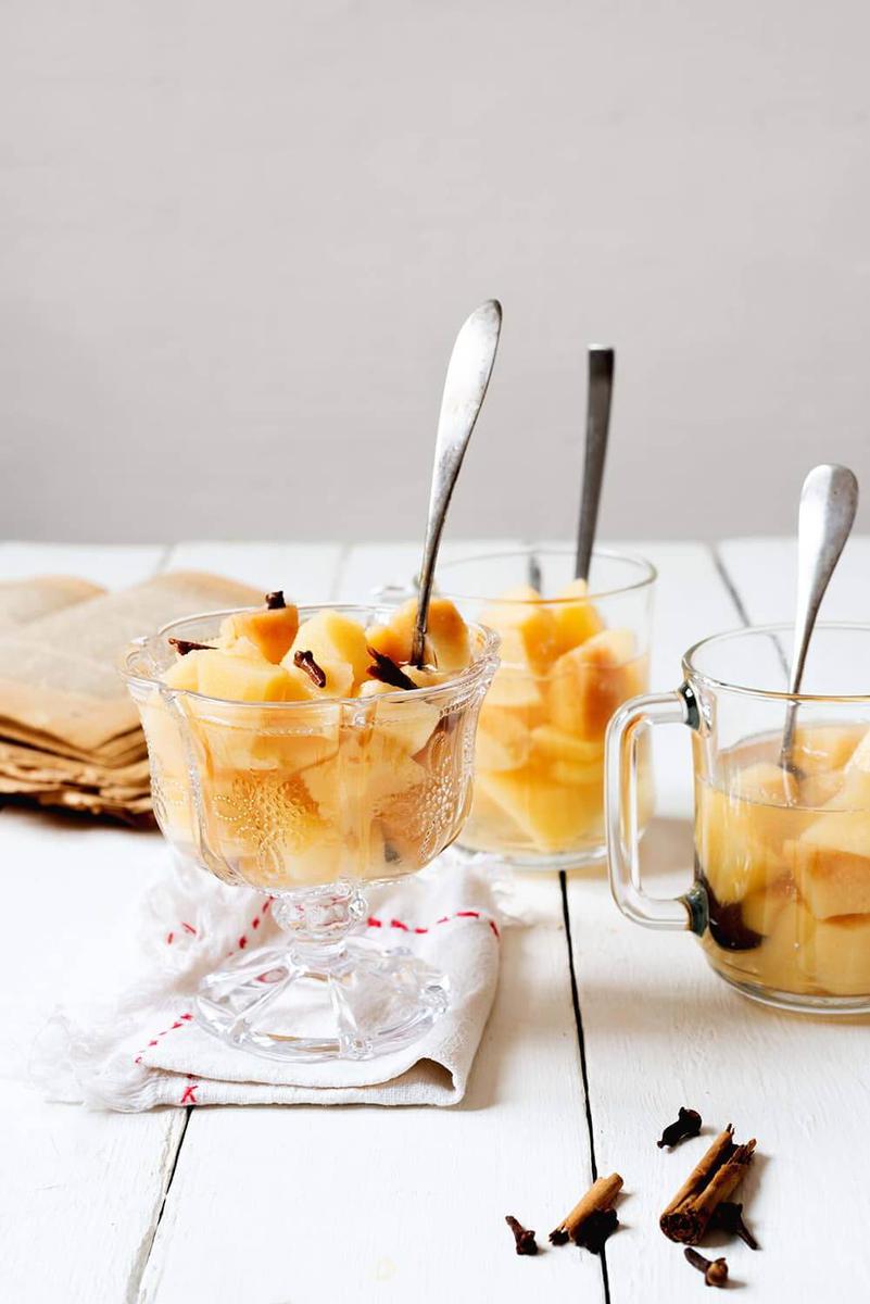 En una mesa blanca están tres copas lleno de compota de membrillo. En cada copa hay una cuchara. Detrás de las copas hay un libro de cocina abierta.