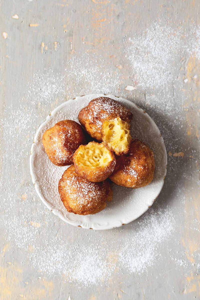En una superficie gris está un plato blanco. En él hay dos buñuelos de viento espolvoreados con azúcar glas.